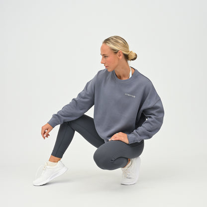 Women’s HYBRYD oversized organic cotton sweatshirt in washed slate grey, with chest logo, styled with dark grey gym leggings.