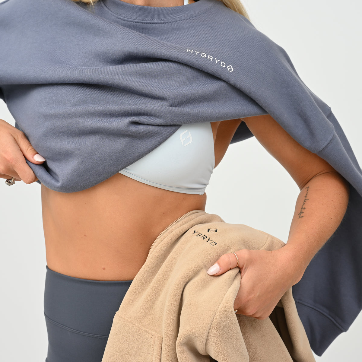 A close-up of a model wearing a slate blue-gray sweatshirt with 'HYBRYD' printed in white on the chest, lifting it to reveal a light gray sports bra. She is also holding a beige sweatshirt with 'HYBRYD' printed on it against a clean white background.