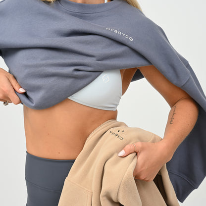 A close-up of a model wearing a slate blue-gray sweatshirt with 'HYBRYD' printed in white on the chest, lifting it to reveal a light gray sports bra. She is also holding a beige sweatshirt with 'HYBRYD' printed on it against a clean white background.