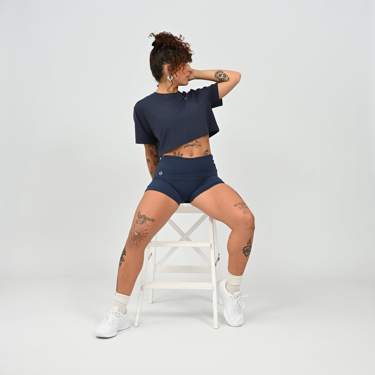 A training tee, oversized, cropped fit in Navy colour. HYBRYD branding on upper left chest. Worn by a gym girl sat on a stool. From activewear brand Hybryd.