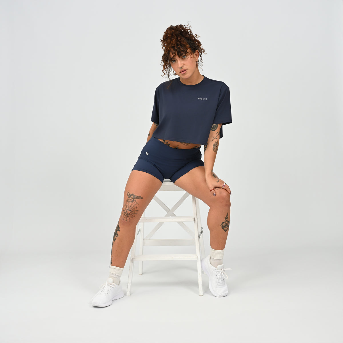 A training tee, oversized, cropped fit in Navy colour. HYBRYD branding on upper left chest. Worn by a gym girl sat on a stool. From activewear brand Hybryd.