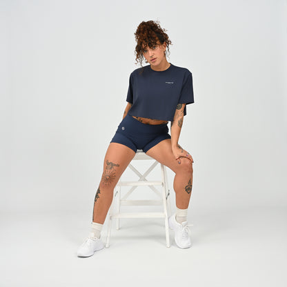 A training tee, oversized, cropped fit in Navy colour. HYBRYD branding on upper left chest. Worn by a gym girl sat on a stool. From activewear brand Hybryd.