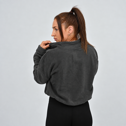 A gym women modeling a dark gray cropped long-sleeve quarter-zip pullover with a white HYBRYD logo on the left chest, paired with black Hybryd leggings, against a solid light gray background.