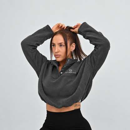 A woman modeling a dark gray cropped long-sleeve quarter-zip pullover with a white HYBRYD logo on the left chest, paired with black Hybryd leggings, against a solid light gray background.