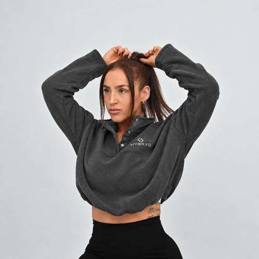 A woman modeling a dark gray cropped long-sleeve quarter-zip pullover with a white HYBRYD logo on the left chest, paired with black Hybryd leggings, against a solid light gray background.