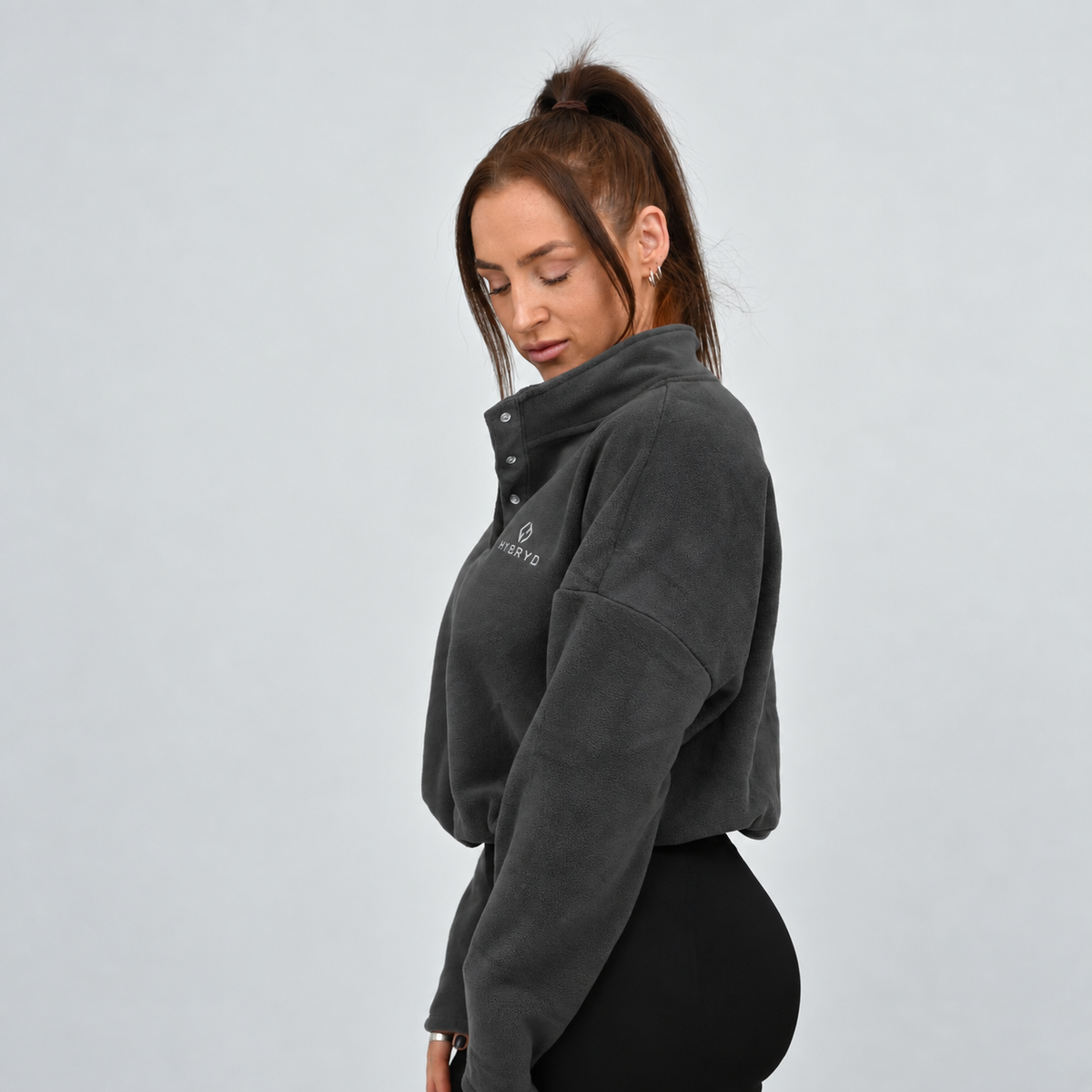 A woman wearing a dark gray cropped fleece pullover with a half-placket and snap buttons, paired with black Hybryd leggings.