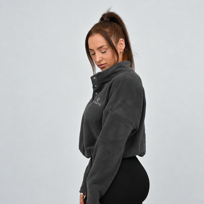 A woman wearing a dark gray cropped fleece pullover with a half-placket and snap buttons, paired with black Hybryd leggings.