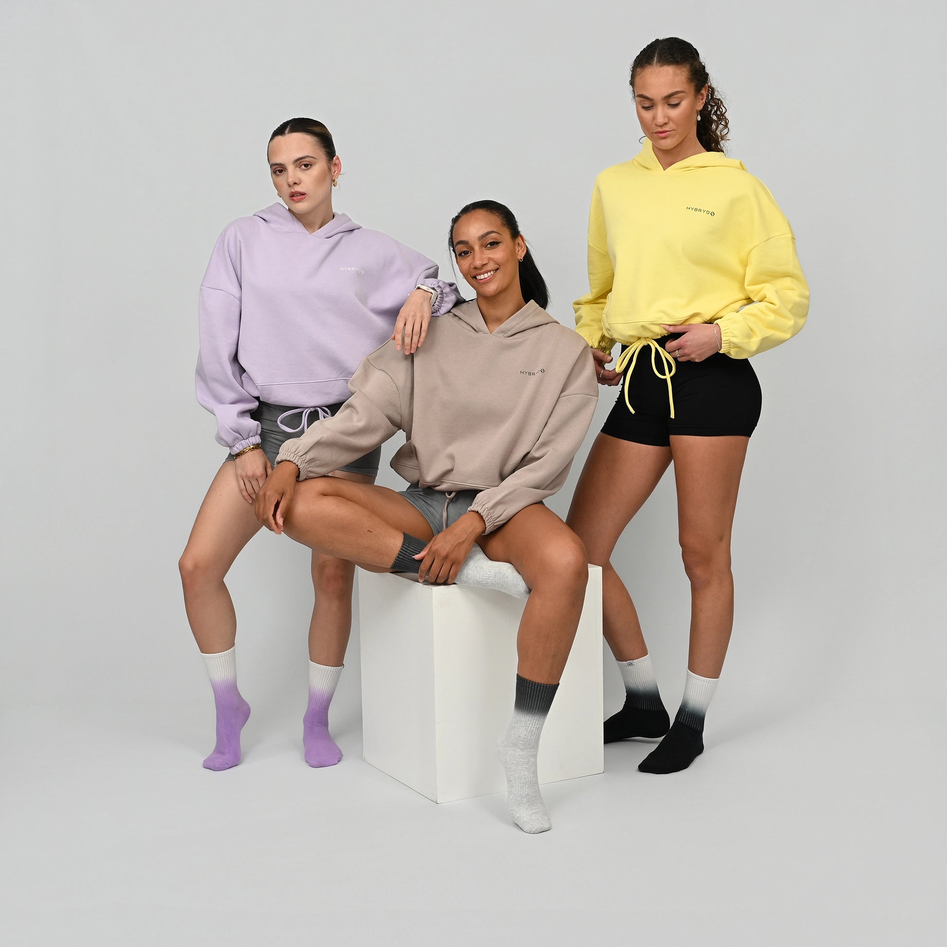 Three women wearing colorful Hybryd hoodies and shorts on a plain background. 