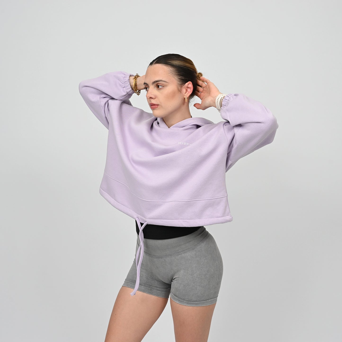 Woman wearing a light purple hoodie from Hybryd and grey booty shorts on a plain background
