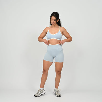 Hyrox athlete wearinga pair of powder blue booty shorts with white piping. High waisted with a cinch bum and contouring. Worn with a matching minimal sports bra. From the brand Hybryd.