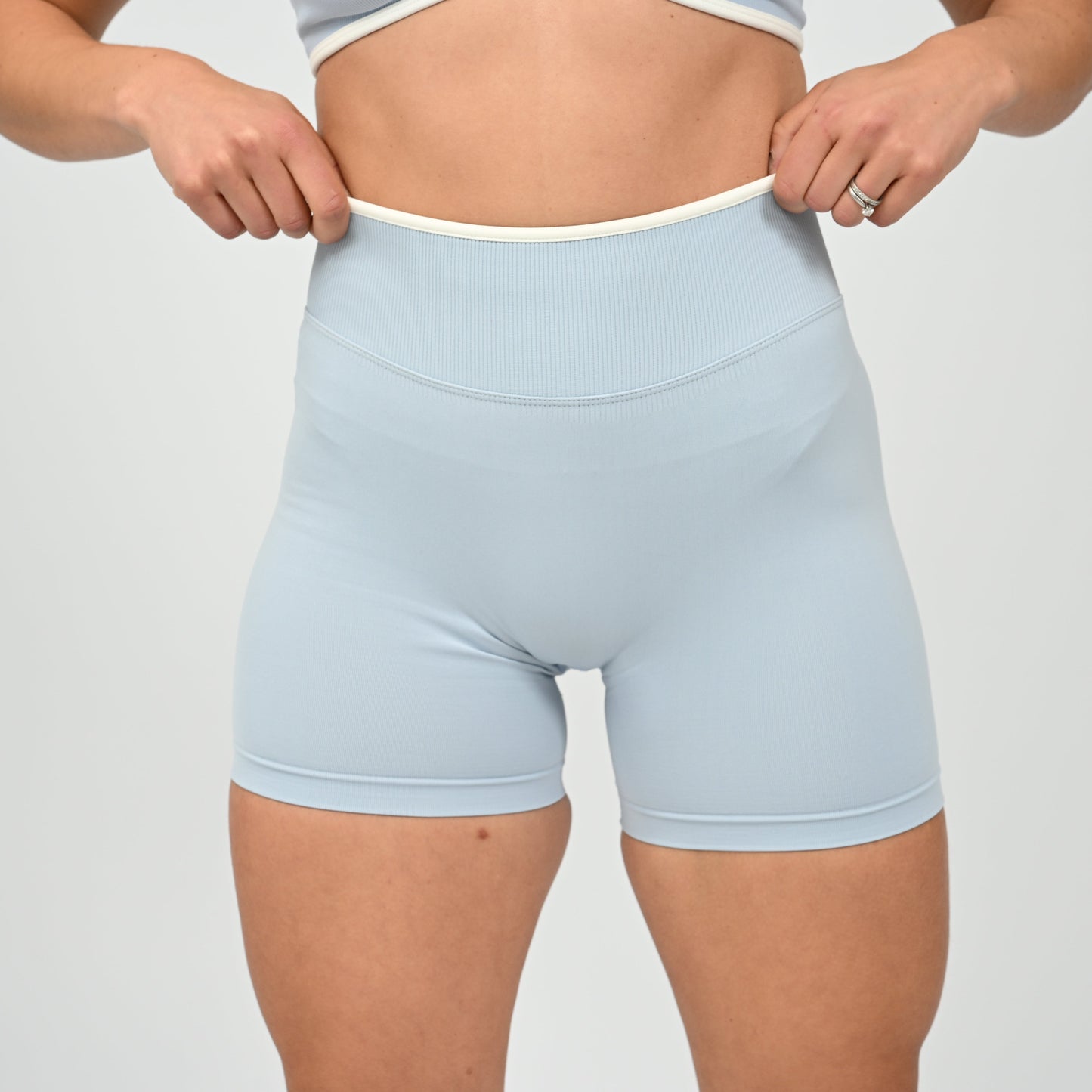 Hyrox athlete wearinga pair of powder blue booty shorts with white piping. High waisted with a cinch bum and contouring. Worn with a matching minimal sports bra. From the brand Hybryd.