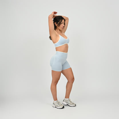 Hyrox athlete wearinga pair of powder blue booty shorts with white piping. High waisted with a cinch bum and contouring. Worn with a matching minimal sports bra. From the brand Hybryd.