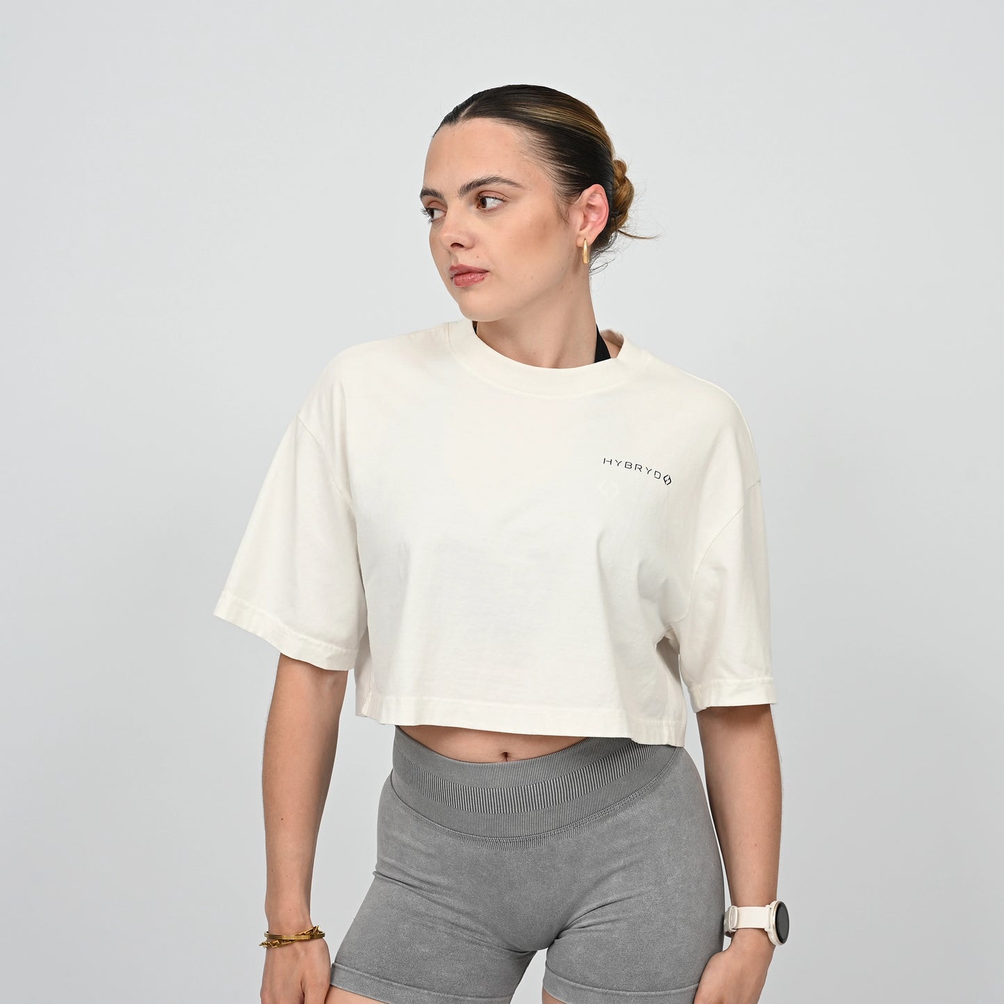 Woman wearing a white crop top and grey gym sorts from the brand Hybryd on a light grey background
