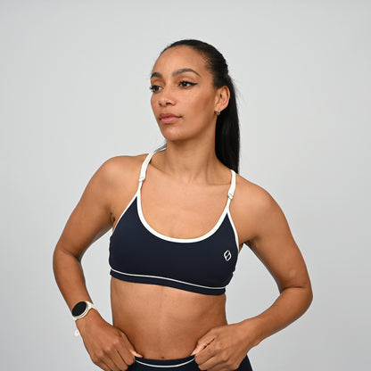 Female crossfit athlete wearing a navy minimal sports bra with white adjustable straps and white piping against a grey background. From activewear brand Hybryd.