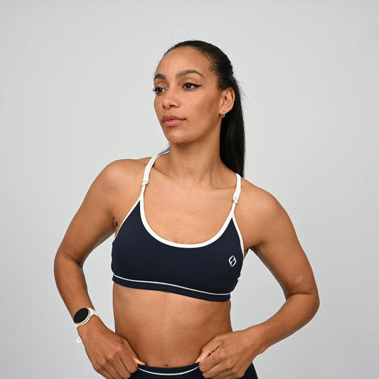 Female crossfit athlete wearing a navy minimal sports bra with white adjustable straps and white piping against a grey background. From activewear brand Hybryd.