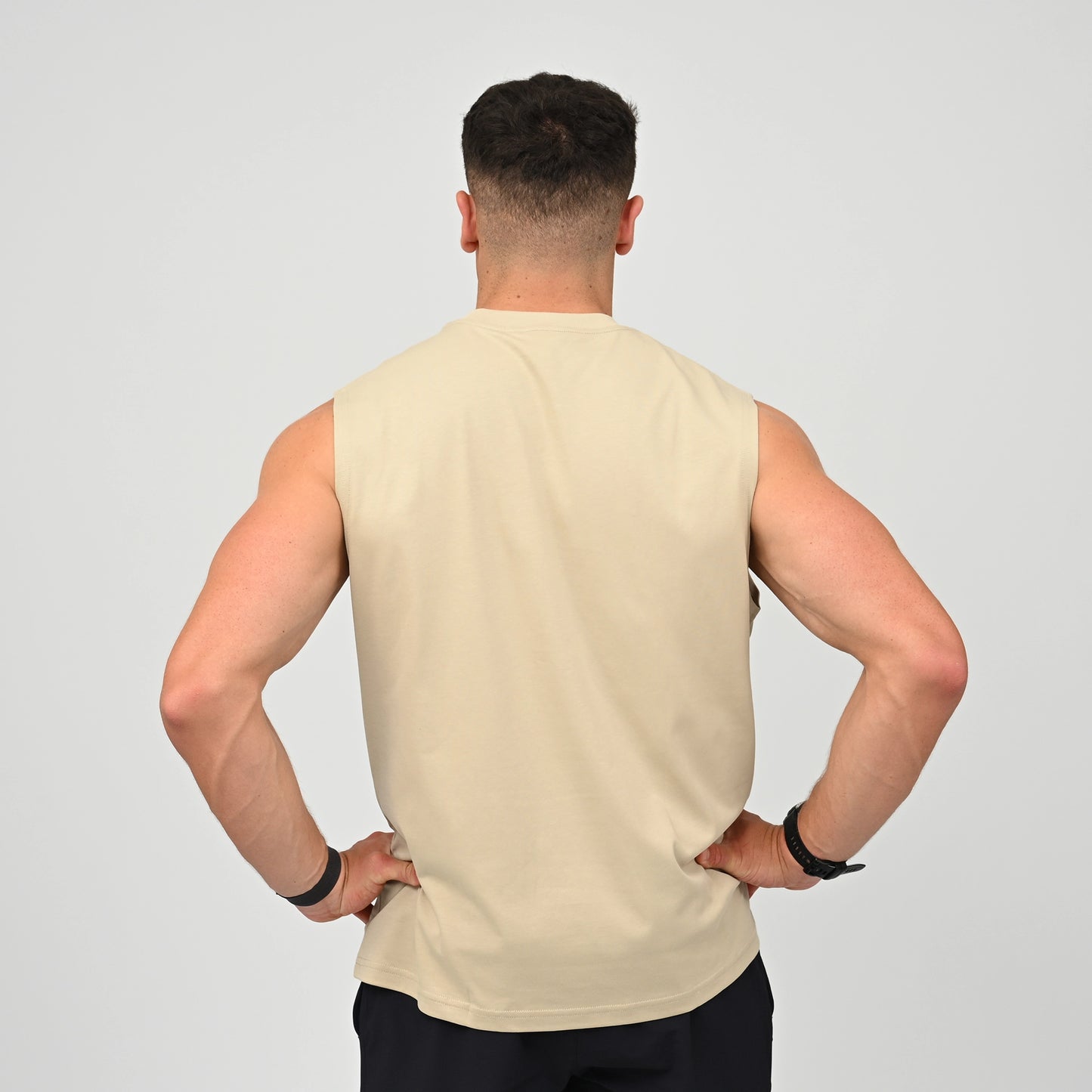 A man viewed from behind wearing a sleeveless beige athletic Hybryd tank top and black pants, standing with hands on hips against a light gray studio background.