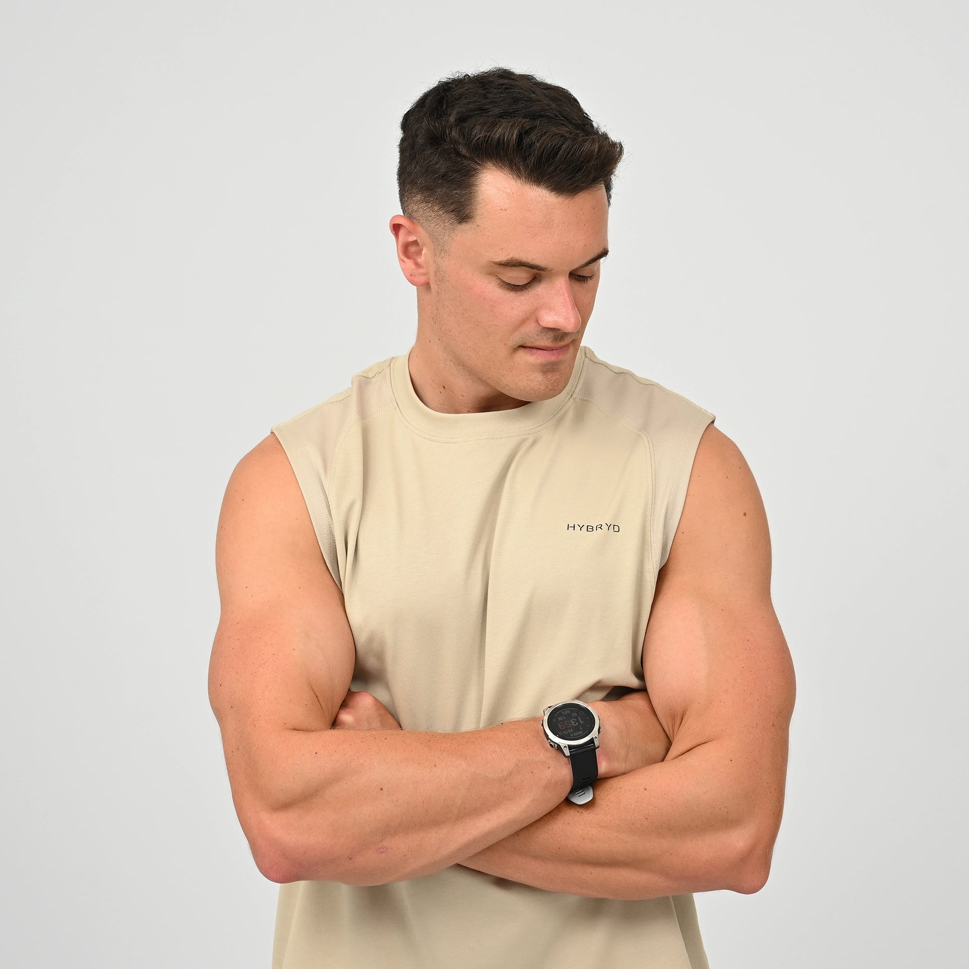 A muscular man wearing a beige sleeveless athletic tank shirt with a small 'HYBRYD' logo on the left chest, paired with a black smartwatch, set against a solid light gray background.