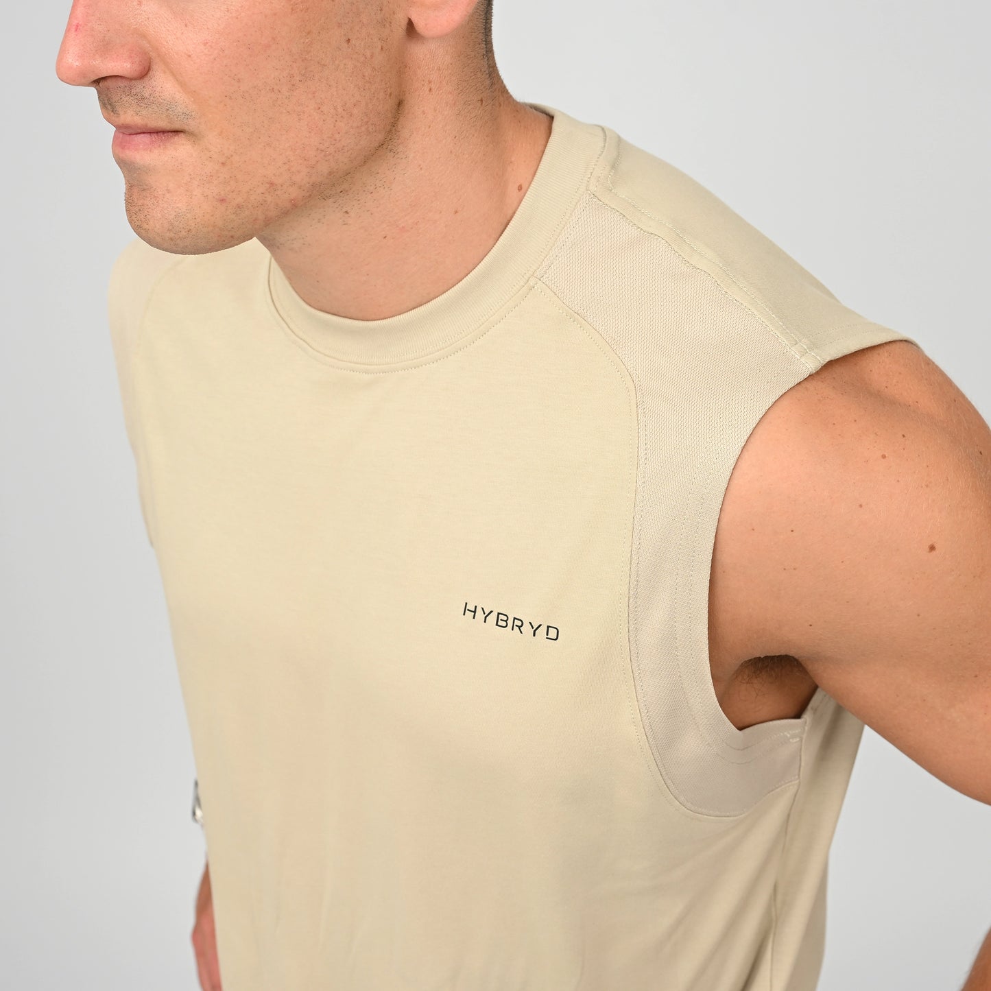 A close-up of a crossfit athlete wearing a light beige sleeveless athletic gym vest with 'HYBRYD' printed on the chest, set against a plain light gray background.