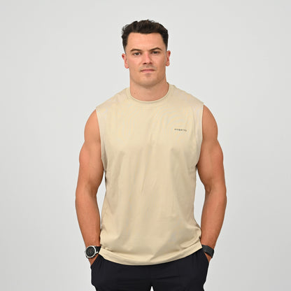 An athlete wearing a beige sleeveless muscle tank top with a small 'Hybryd' logo on the left chest, paired with black gym shorts, standing against a light gray studio background.