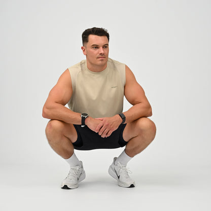 A muscular man in a crouching pose wearing a beige sleeveless athletic shirt, black athletic shorts, white socks, and white Nike running shoes against a light gray studio background.