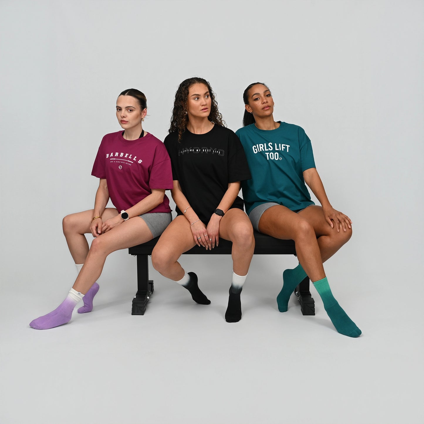 Three women sitting on a bench wearing t-shirts with text and colorful socks. From gymwear brnad Hybryd.