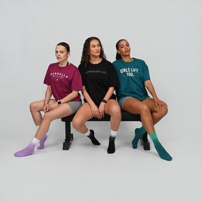 Three women sitting on a bench wearing t-shirts with text and colorful socks. From gymwear brnad Hybryd.
