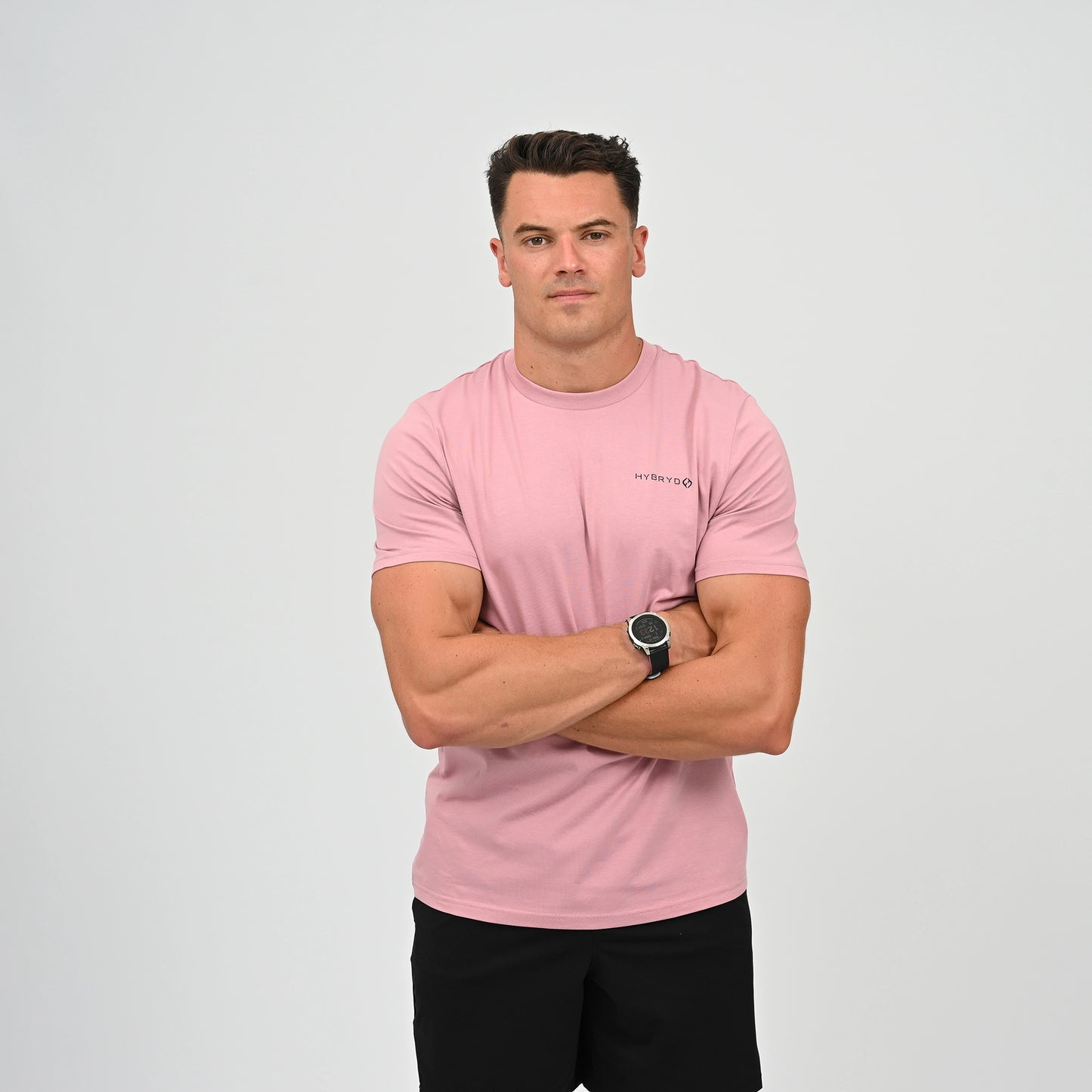 A Crossfitter wearing a light pink crew-neck t-shirt with a small black Hybryd logo on the left chest, paired with black pants, standing against a light gray studio background. From Hybryd.