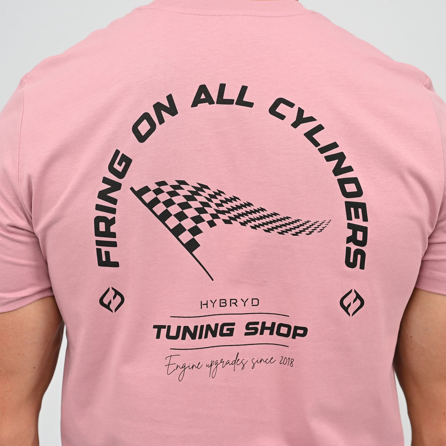 A Hyrox athlete viewed from behind wearing a light pink t-shirt with a black graphic print on the back. The graphic features the text 'FIRING ON ALL CYLINDERS' in an arch, a checkered flag motif, and 'TUNING SHOP' tag line. From gymwear brand Hybryd.