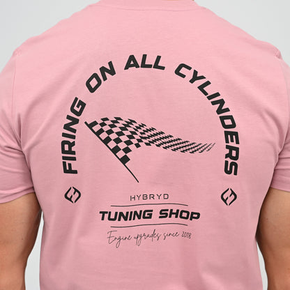 A Hyrox athlete viewed from behind wearing a light pink t-shirt with a black graphic print on the back. The graphic features the text 'FIRING ON ALL CYLINDERS' in an arch, a checkered flag motif, and 'TUNING SHOP' tag line. From gymwear brand Hybryd.