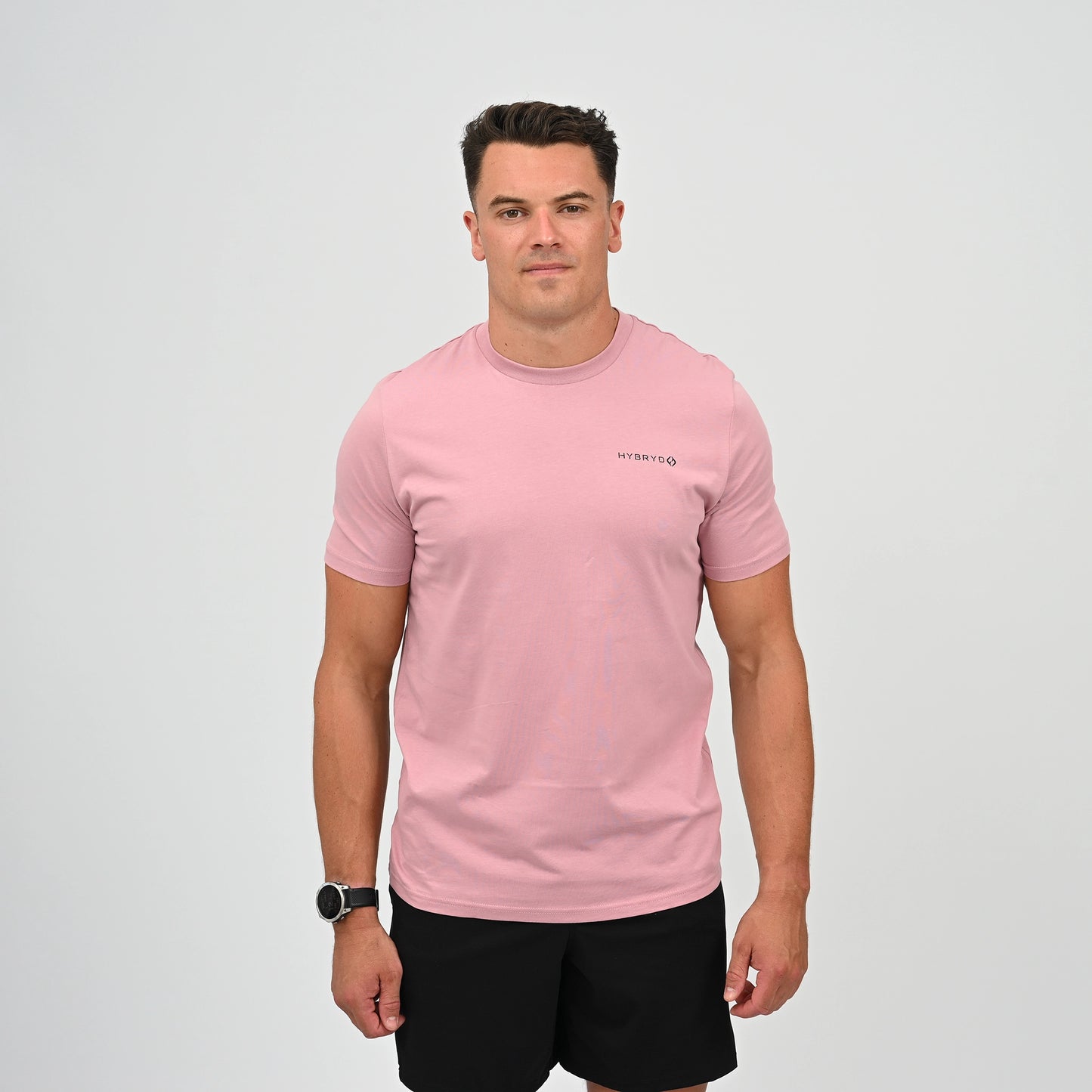 A man wearing a light pink crew-neck t-shirt with a small black Hybryd logo on the left chest, paired with black athletic shorts, standing against a light gray studio background. From Gymwear brand Hybryd.