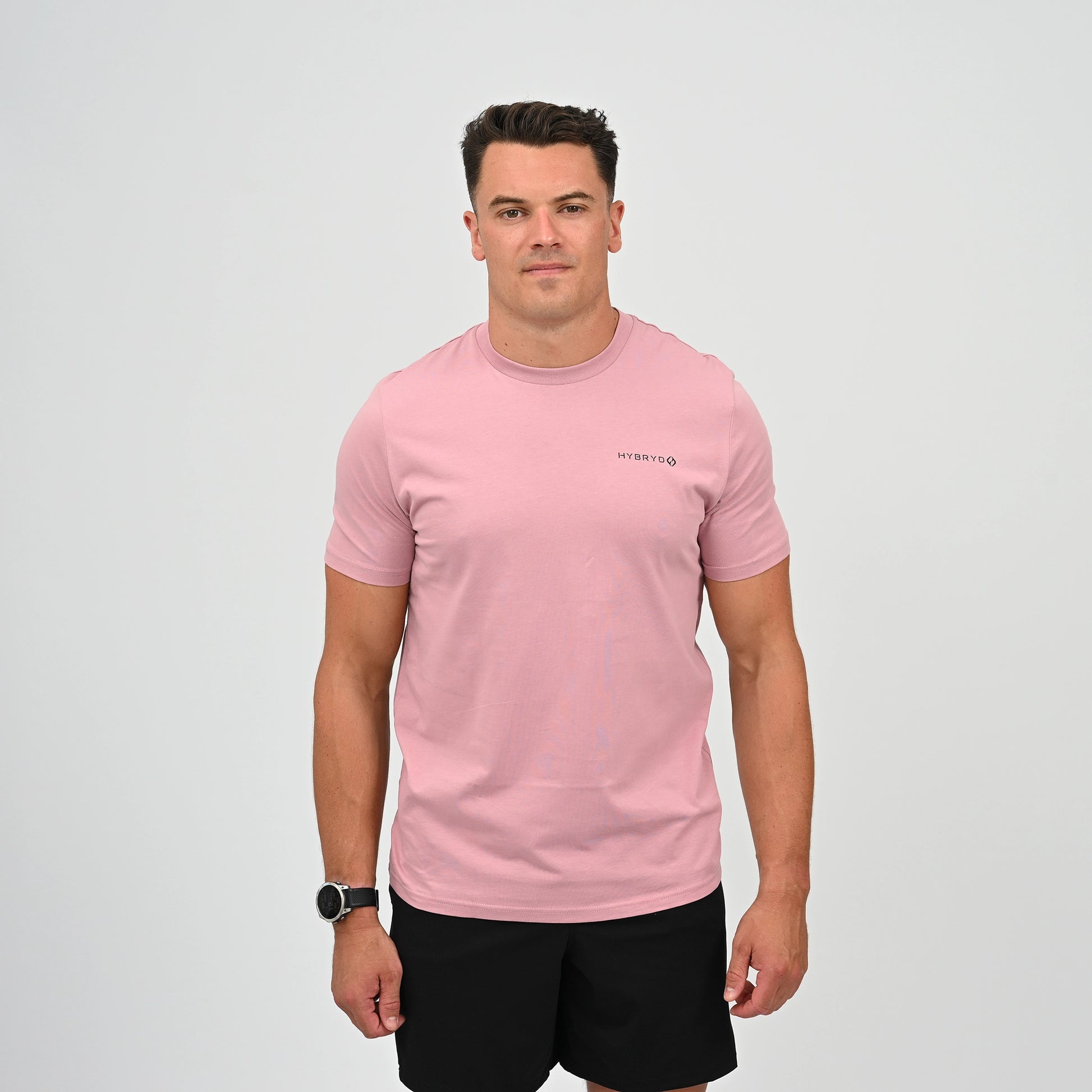 A man wearing a light pink crew-neck t-shirt with a small black Hybryd logo on the left chest, paired with black athletic shorts, standing against a light gray studio background. From Gymwear brand Hybryd.