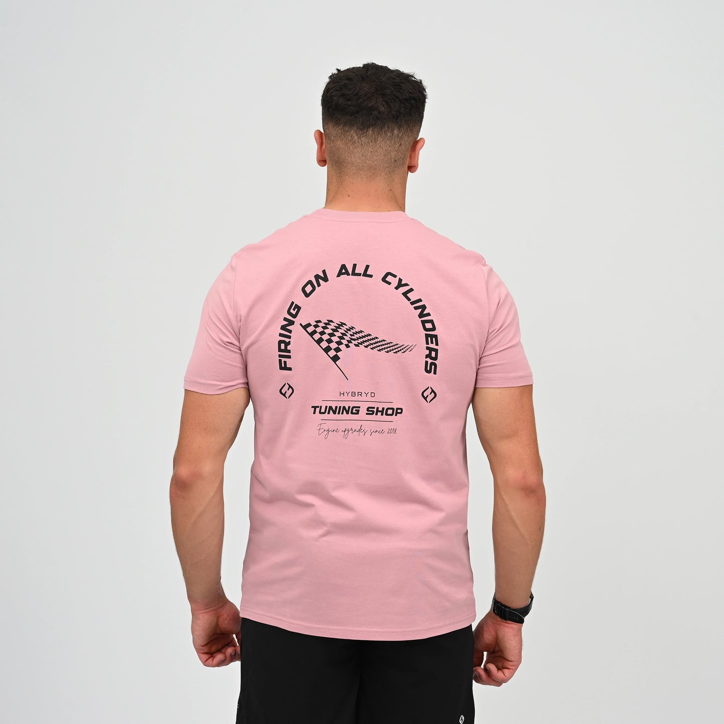 A Hyrox athlete viewed from behind wearing a light pink t-shirt with a black graphic print on the back. The graphic features the text 'FIRING ON ALL CYLINDERS' in an arch, a checkered flag motif, and 'TUNING SHOP' tag line. From gymwear brand Hybryd.