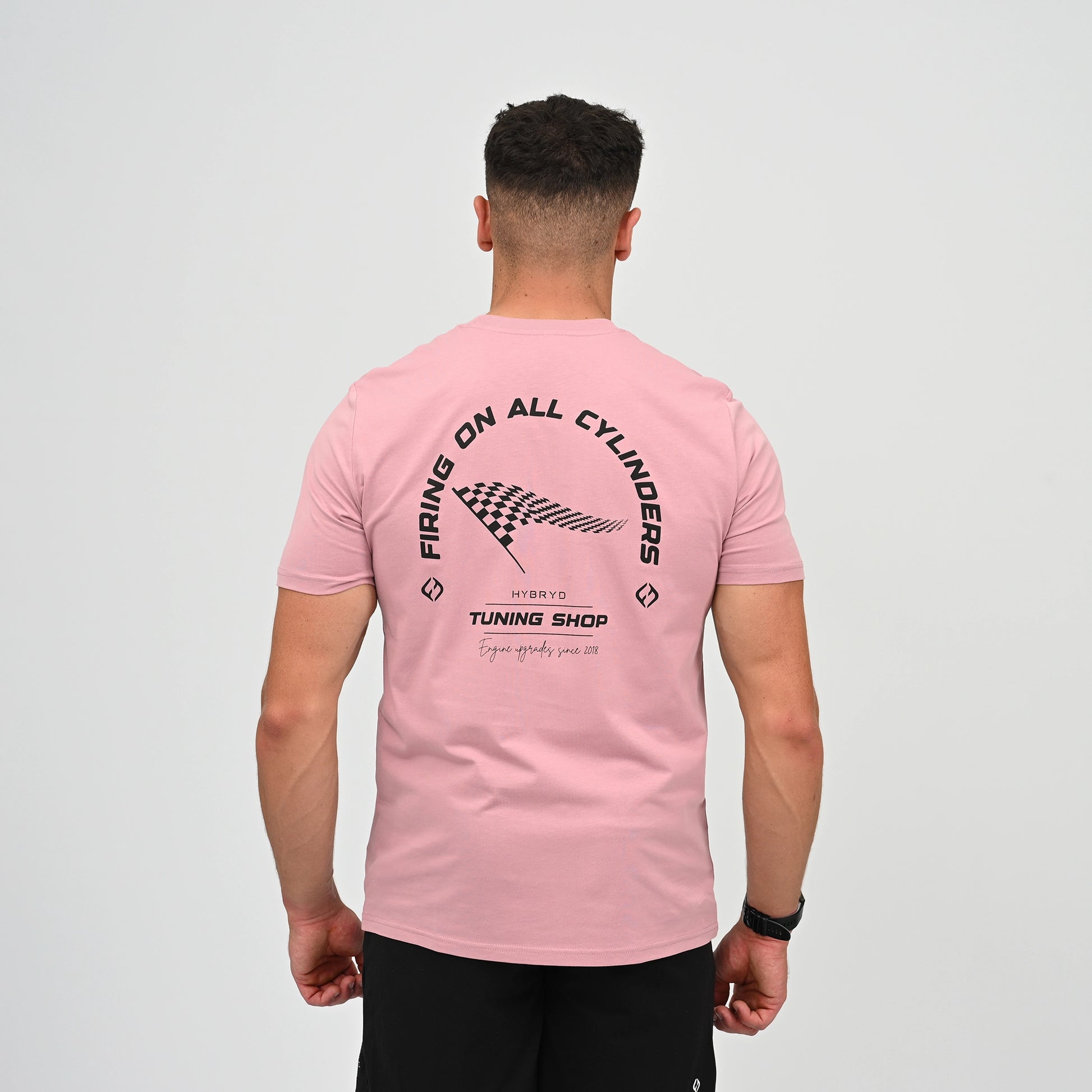 A Hyrox athlete viewed from behind wearing a light pink t-shirt with a black graphic print on the back. The graphic features the text 'FIRING ON ALL CYLINDERS' in an arch, a checkered flag motif, and 'TUNING SHOP' tag line. From gymwear brand Hybryd.
