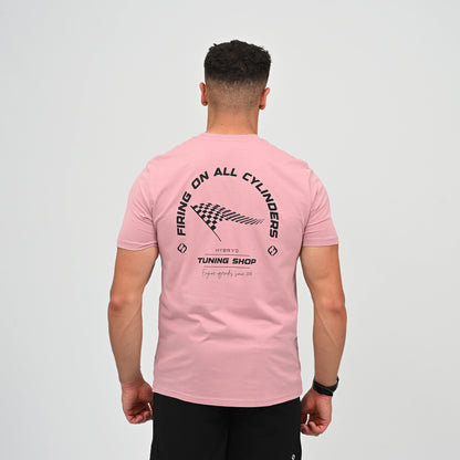 A Hyrox athlete viewed from behind wearing a light pink t-shirt with a black graphic print on the back. The graphic features the text 'FIRING ON ALL CYLINDERS' in an arch, a checkered flag motif, and 'TUNING SHOP' tag line. From gymwear brand Hybryd.