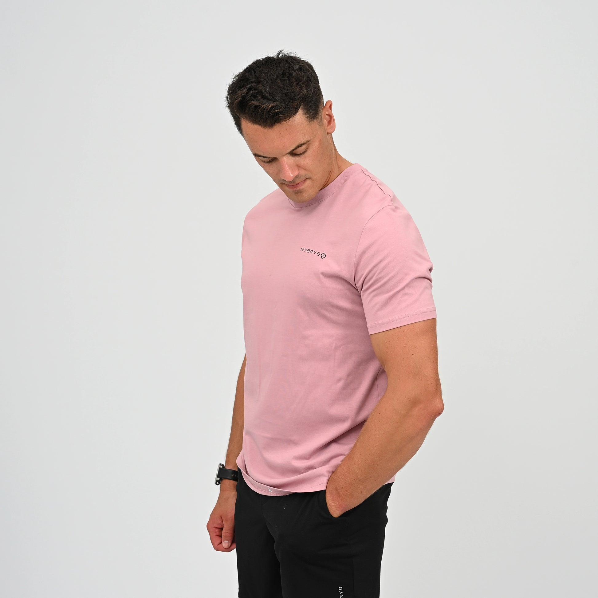 A Crossfitter wearing a light pink crew-neck t-shirt with a small black Hybryd logo on the left chest, paired with black pants, standing against a light gray studio background. From Hybryd.