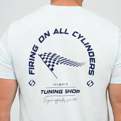 A Hyrox athlete viewed from behind wearing a light blue t-shirt with a navy graphic print on the back. The graphic features the text 'FIRING ON ALL CYLINDERS' in an arch, a checkered flag motif, and 'TUNING SHOP' tag line. From gymwear brand Hybryd.