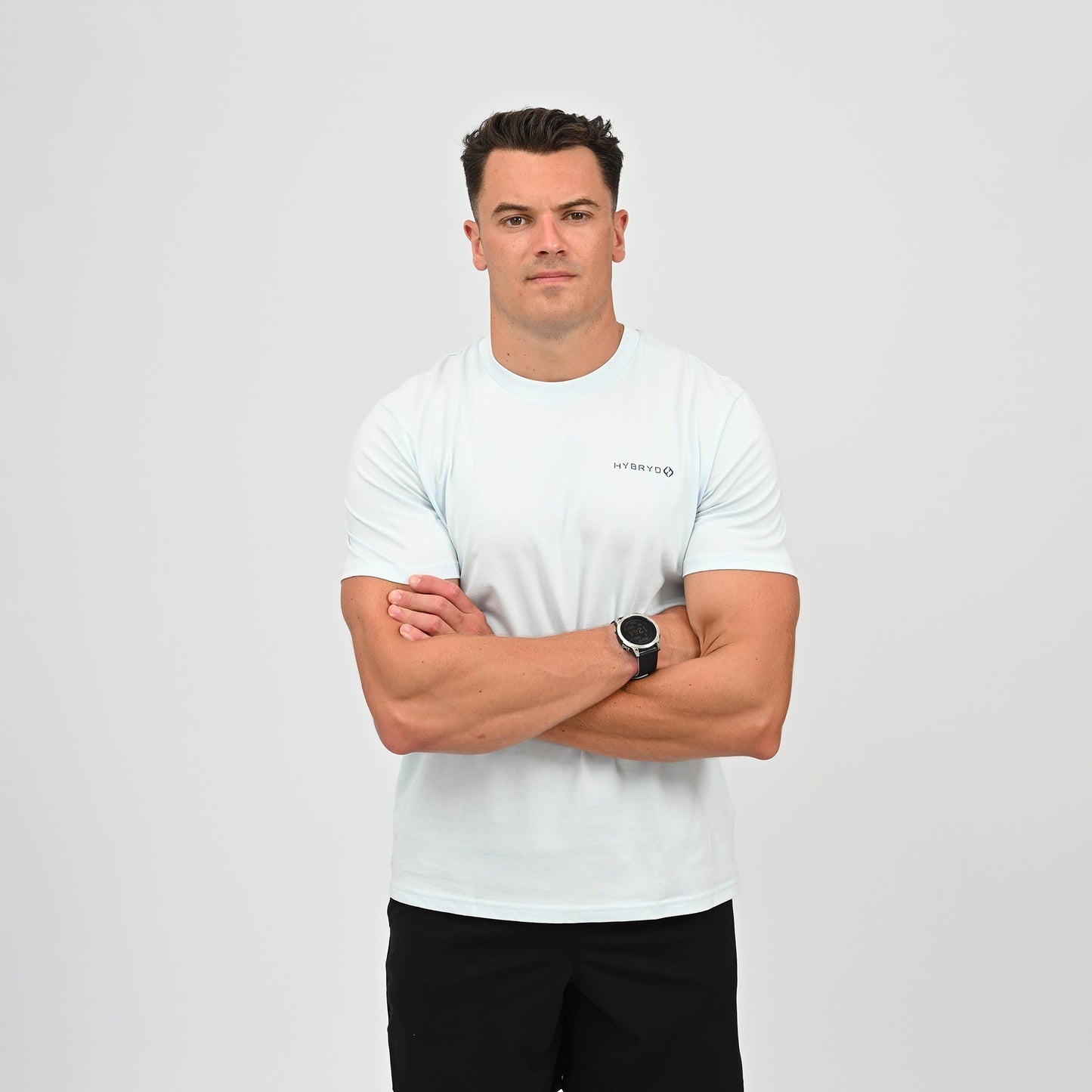 A man wearing a light blue crew-neck t-shirt with a small black Hybryd logo on the left chest, paired with black athletic shorts, standing against a light gray studio background. From Gymwear brand Hybryd.
