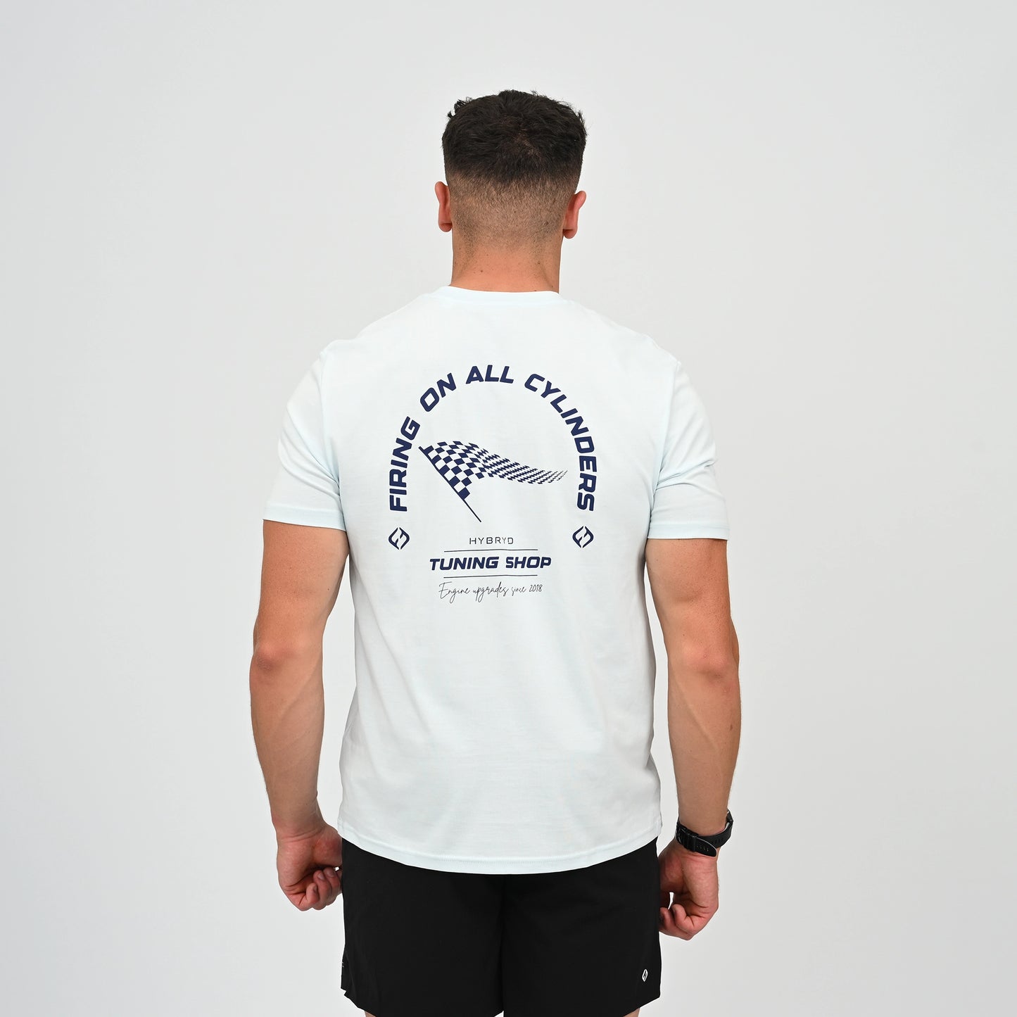 A Hyrox athlete viewed from behind wearing a light blue t-shirt with a black graphic print on the back. The graphic features the text 'FIRING ON ALL CYLINDERS' in an arch, a checkered flag motif, and 'TUNING SHOP' tag line. From gymwear brand Hybryd.