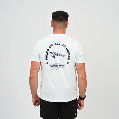 A Hyrox athlete viewed from behind wearing a light blue t-shirt with a black graphic print on the back. The graphic features the text 'FIRING ON ALL CYLINDERS' in an arch, a checkered flag motif, and 'TUNING SHOP' tag line. From gymwear brand Hybryd.