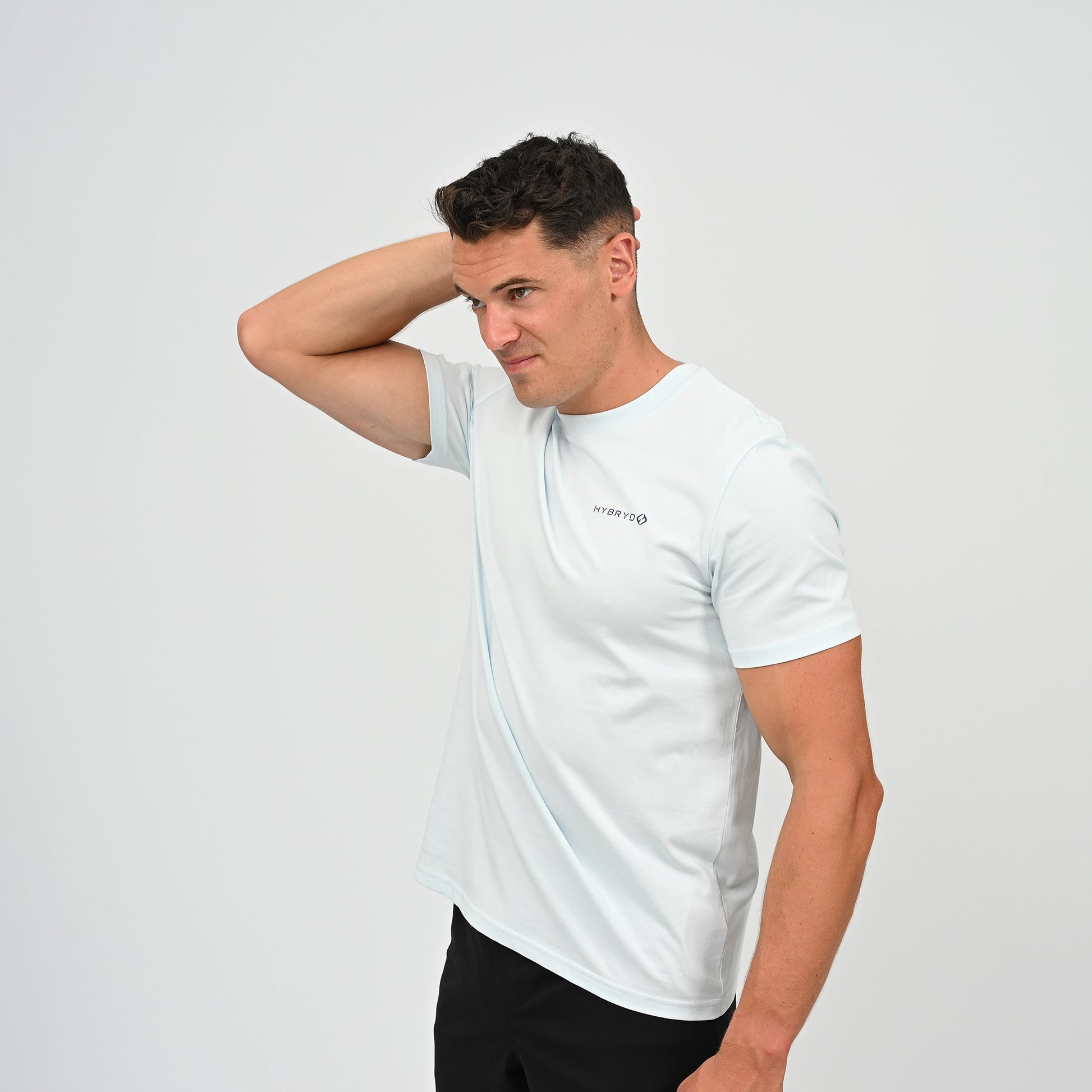 A man wearing a light blue crew-neck t-shirt with a small black Hybryd logo on the left chest, paired with black athletic shorts, standing against a light gray studio background. From Gymwear brand Hybryd.