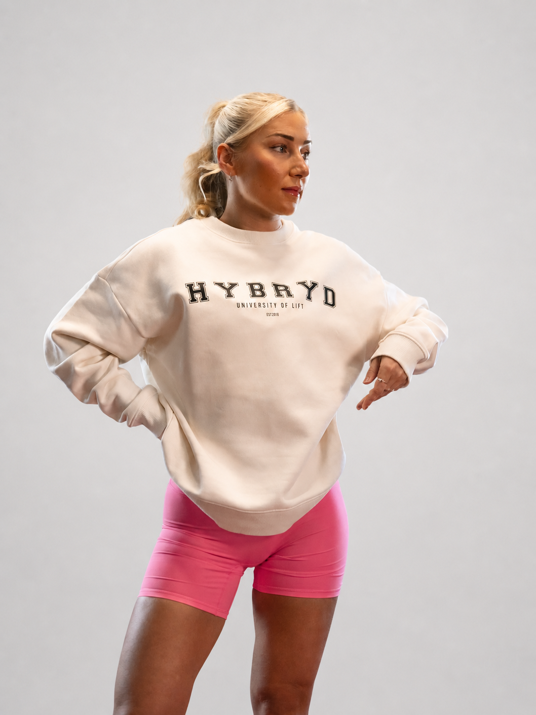 Person wearing a cream sweatshirt with 'HYBRYD' text in a varsity style. With pink shorts on a grey background