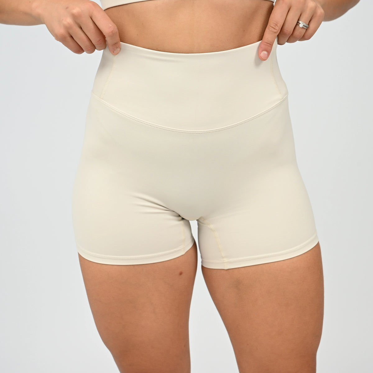 A close-up of a model wearing high-waisted Hybryd shorts in a light cream color, with their hands pulling slightly at the waistband to demonstrate stretch. 