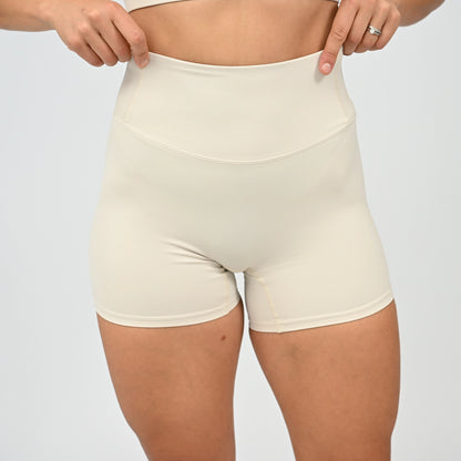 A close-up of a model wearing high-waisted Hybryd shorts in a light cream color, with their hands pulling slightly at the waistband to demonstrate stretch. 