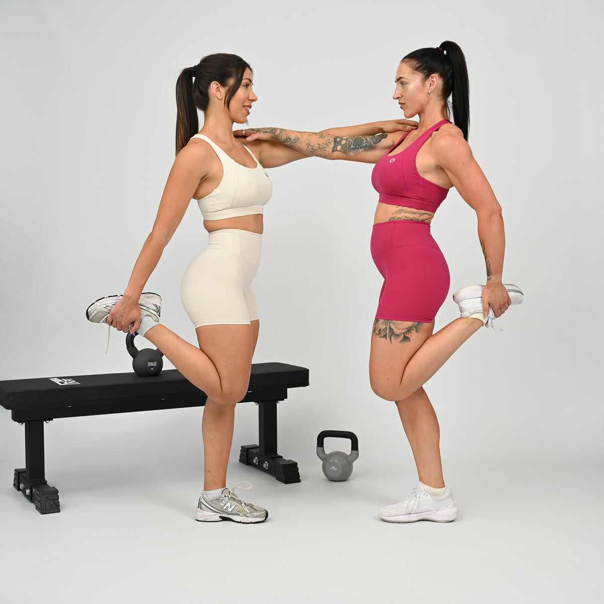 Two women in matching athletic wear from Hybryd, perform a stretching exercise in a studio setting. One wears a cream Hybryd sports bra and shorts, and the other wears a bright pink sports bra and shorts.