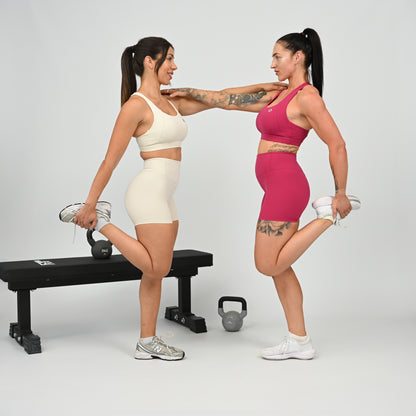 Two women in matching athletic wear from Hybryd, perform a stretching exercise in a studio setting. One wears a cream Hybryd sports bra and shorts, and the other wears a bright pink sports bra and shorts.