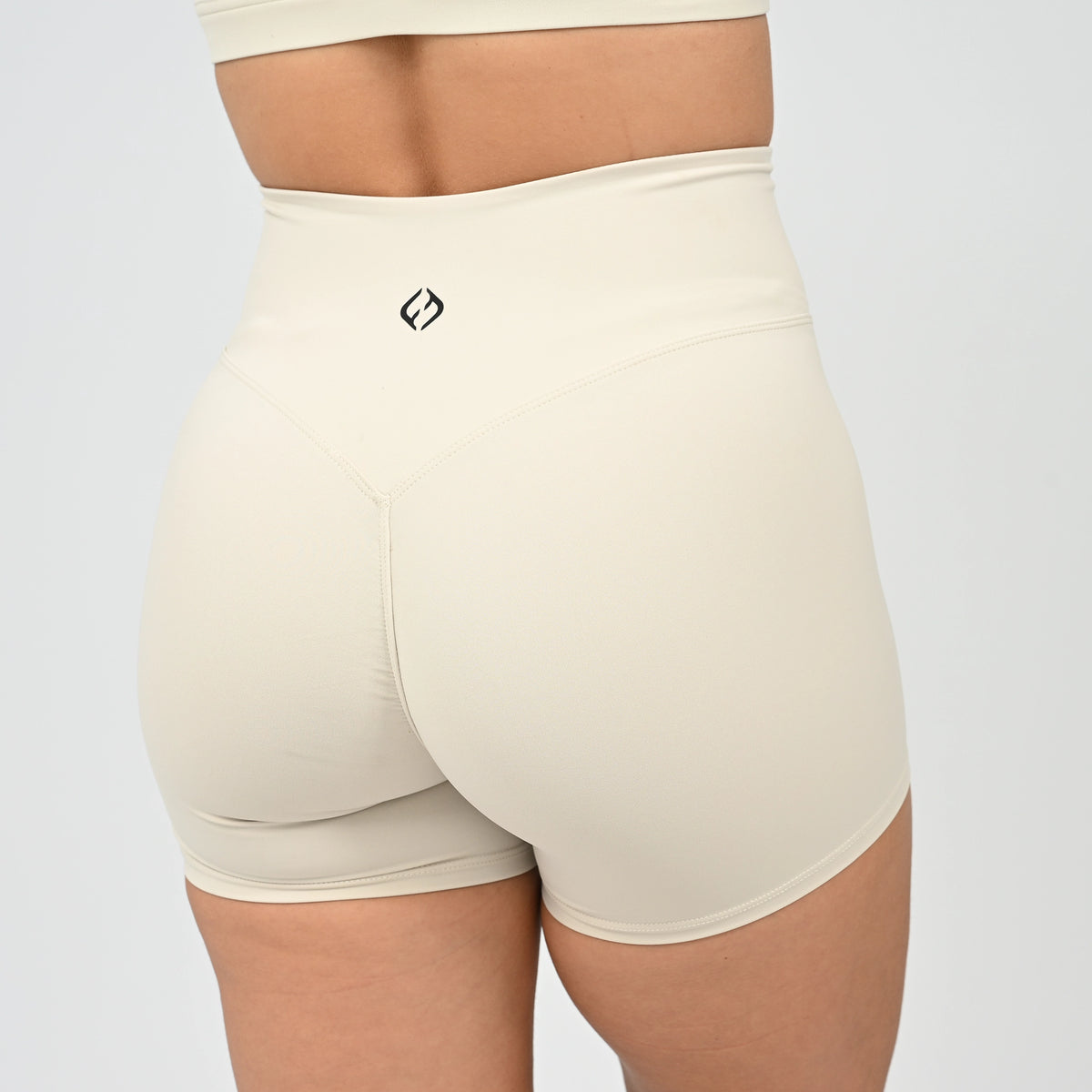 Rear view of a person wearing high-waisted beige athletic shorts with a small black Hybryd logo on the back waistband, slight cinch in the bum. Against a light gray studio background.