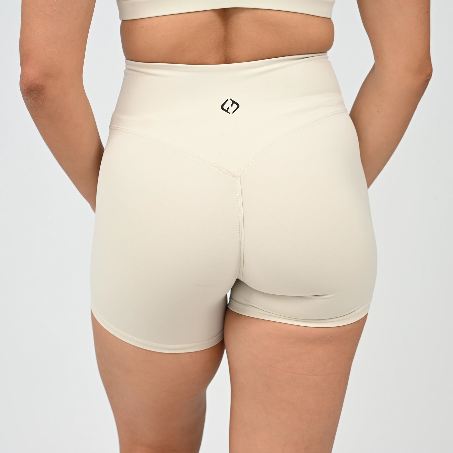 Rear view of a Hyrox model wearing high-waisted booty shorts in a light cream color, paired with a matching sports bra against a light gray studio background.