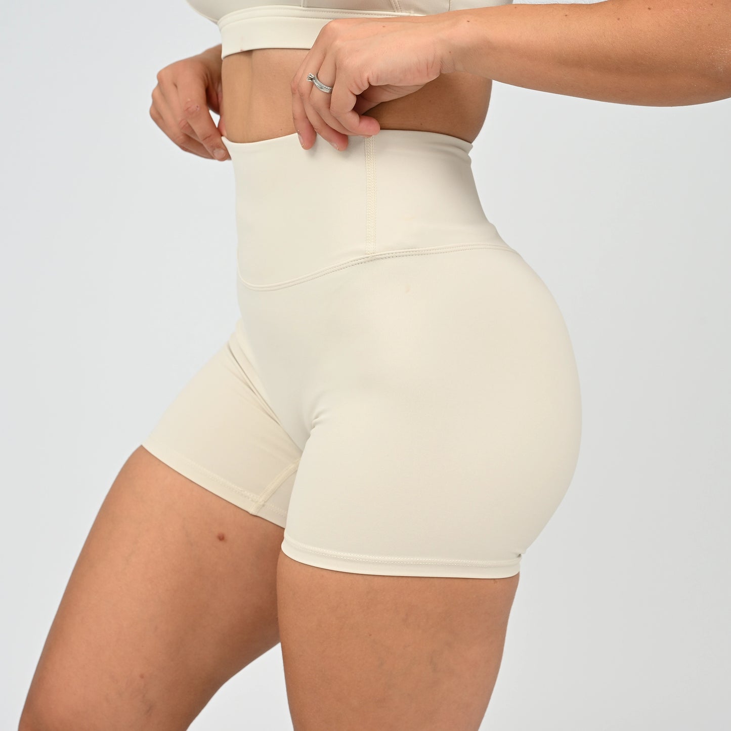 A close-up of a model wearing high-waisted, form-fitting athletic shorts in a light cream or off-white color, paired with a matching crop top against a plain light gray background. From activewear brand Hybryd.