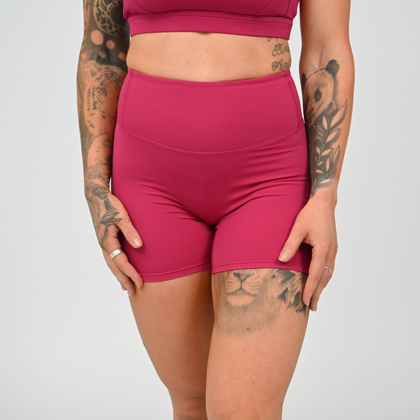A close-up of a model wearing high-waisted athletic shorts in a vibrant magenta pink, paired with a matching sports bra. The model has visible tattoos on both arms and thigh, and is photographed against a neutral light gray studio background. From Hybryd gymwear.