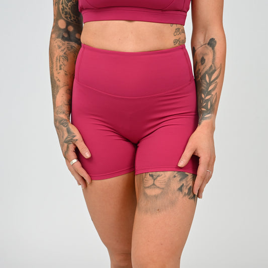 A close-up of a model wearing high-waisted athletic shorts in a vibrant magenta pink, paired with a matching sports bra. The model has visible tattoos on both arms and thigh, and is photographed against a neutral light gray studio background. From Hybryd gymwear.